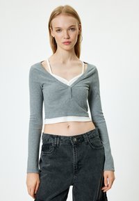 Grau geschnittenes Langarmoberteil mit V-Ausschnitt, ausgestattet mit drei Knöpfen vorne. Kombiniert mit dunkelhohen Hosen. Weißes Trägershirt darunter sichtbar.