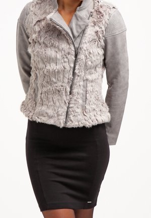 Femme portant une veste en fausse fourrure gris clair et en suède avec une fermeture éclair asymétrique, associée à une jupe crayon noire ajustée.