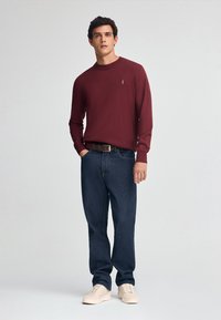 Burgundowy sweter z długim rękawem i okrągłym dekoltem, w połączeniu z niebieskimi dżinsami i kremowymi trampkami. Prosty detal logo na swetrze.