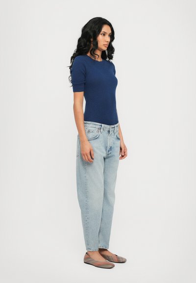Navy ribbed knit top, light blue relaxed-fit jeans, and multicolored striped flats. Model stands in profile against a plain background.