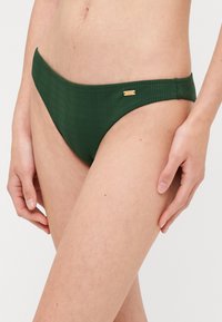 Gröna ribbade bikiniunderbyxor med en slät midjeband och en liten guldfärgad accenttag på sidan. Tyget har ett mjukt, texturerat utseende.