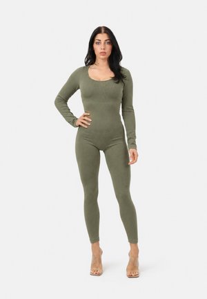 Femme debout, main sur la hanche, portant une combinaison moulante côtelée vert olive à manches longues et des sandales à talons transparents sur fond blanc.
