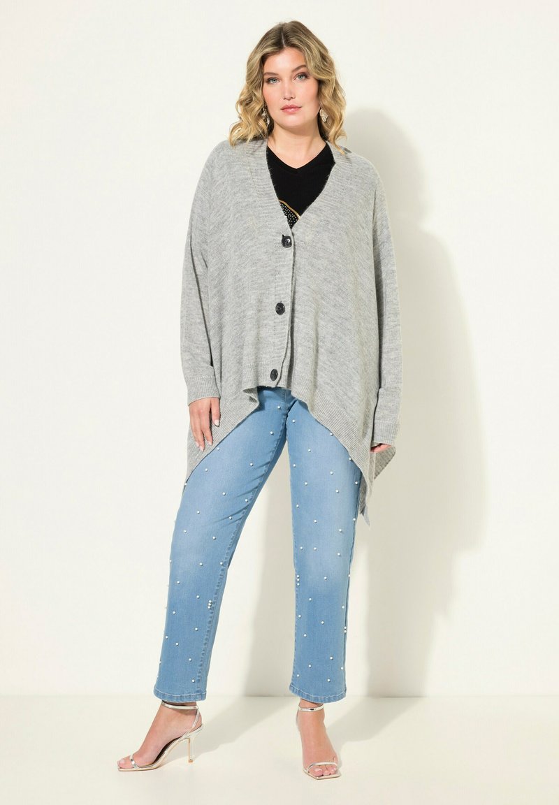 Cardigan grigio a maglia con scollo a V e bottoni neri, abbinato a jeans azzurri chiari con decorazioni in perle e sandali con tacco trasparente.