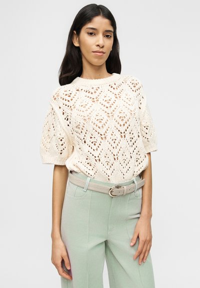 Γυ ναίκα που φοράει κρεμ μακρυμάνικη πλεκτή μπλούζα και ψηλόμεσο, ανοιχτό πράσινο παντελόνι με μπεζ ζώνη, στέκεται μπροστά σε απλό φόντο.