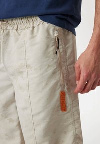 Light beige shorts with a subtle marbled pattern, elastic waistband, side zip pockets, and a prominent orange "GUESS" logo.