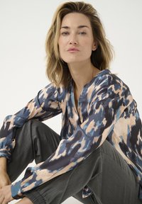 Langærmet bluse i et blåt, beige og sort abstrakt mønster med V-udskæring. Stoffet er glat, kombineret med grå strukturerede bukser.