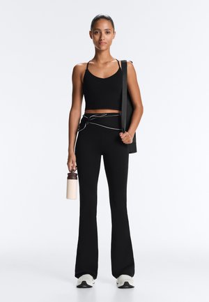 Ženska v črni skrajšani majici brez rokavov in razširjenih pajkicah drži plastenko vode in torbo za jogo, stoji pred enostavnim belim ozadjem.