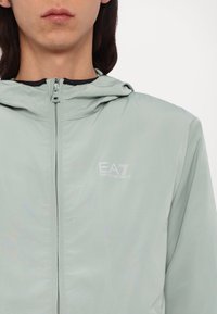 Lys grøn zip-up jakke med hætte, glat tekstur, med EA7-logo i hvid. Minimalistisk design, ingen synlige mønstre eller accenter.