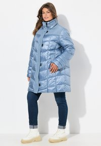 Abrigo puffer azul claro con cuello alto, diseño acolchado, botones a presión y bolsillos laterales. Llevado sobre jeans oscuros y botas blancas gruesas.