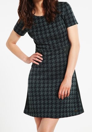 Kvinde iført kortærmet, knælængde kjole med sort og mørkegråt hundetandsmønster, detalje med knapper i siden, poserer med den ene hånd på hoften.
