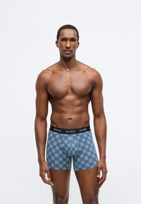 Cuecas boxer com padrão azul apresentando o logótipo "HUGO". A cintura é preta e o tecido parece liso e elástico.