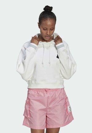 Sudadera blanca recortada con cordones y rayas grises en las mangas, combinada con pantalones cortos de nailon rosas que tienen bolsillos y una etiqueta de marca.