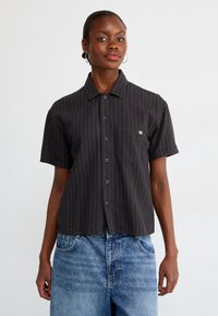 Kortermet svart skjorte med knapper og vertikale gule striper, enkelt brystlomme, og en avslappet passform, kombinert med lyseblå denimbukser.