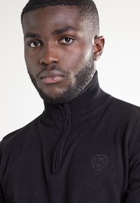 Czarny sweter z zamkiem przy szyi wykonany z miękkiego materiału. Posiada prążkowany kołnierz i subtelne logo. Dopasowany fason z długimi rękawami.