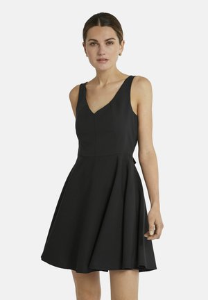 Frau in einem ärmellosen schwarzen A-Linien-Kleid mit V-Ausschnitt, stehend vor einem schlichten hellen Hintergrund.