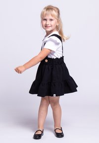 Młoda dziewczynka z blond włosami w kucyku, ubrana w białą koszulę i czarną sukienkę na ramiączkach, uśmiechająca się i stojąca na jasnym tle.