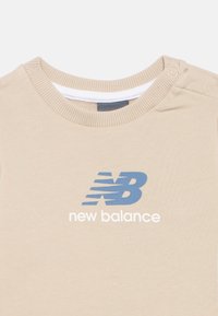 Maglietta beige New Balance con logo blu "NB" e testo bianco "new balance" sotto il collo rotondo e bottoni a pressione sulle spalle.
