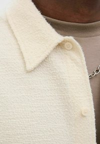Chaqueta de crema texturizada con un cuello tipo solapa, tres botones y una suave tela de punto que cubre una camisa marrón claro debajo.