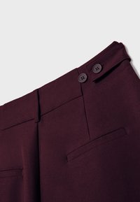 Pantalon ajusté bordeaux foncé à taille haute, avec deux boutons, passants pour ceinture, et deux poches passepoilées à l'arrière sur un fond clair.