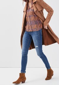 Manteau trench en daim marron porté sur une chemise à motifs multicolores, associé à un jean skinny bleu et des bottines marron à bout pointu.