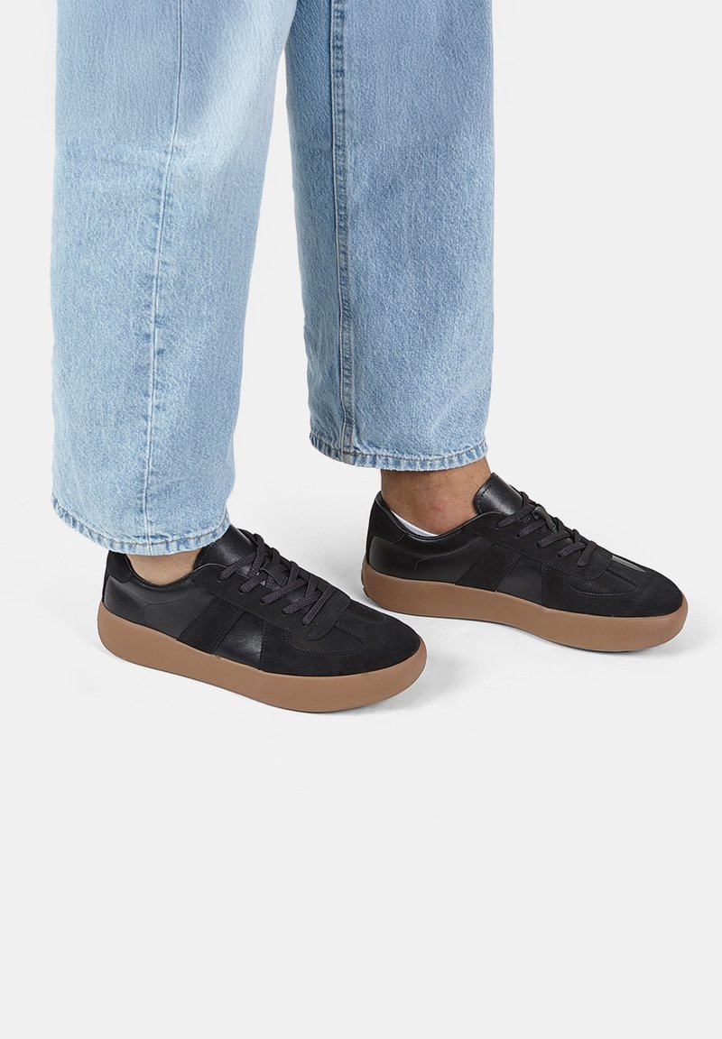 Svarta sneakers i mocka och läder, med en gummisul i brun färg, rund tå och platta snörning. Bärs med ljusblå jeans.