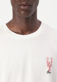 Tricou de culoare crem, realizat din bumbac, cu un grafic de homar roșu și text negru pe piept. Design simplu cu gât rotund.