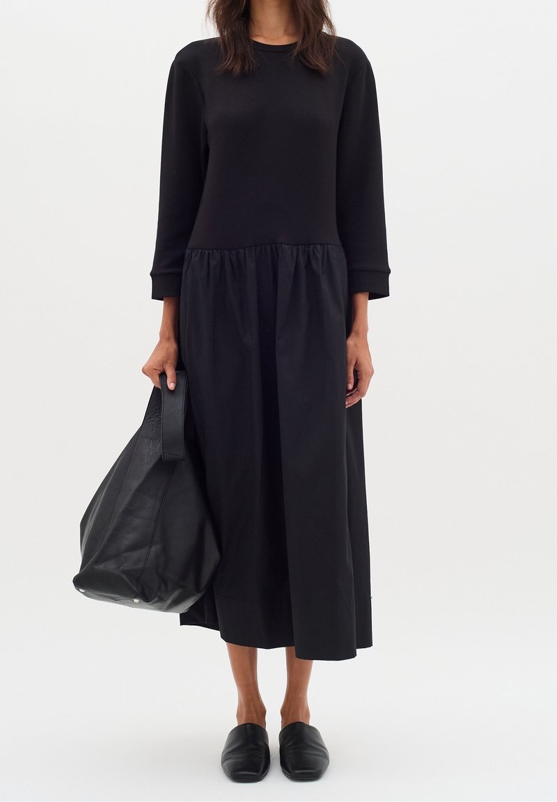 Femme portant une longue robe noire aux manches trois-quarts, tenant un grand sac en cuir noir et portant des chaussures noires sans lacets.