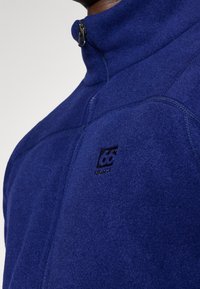 Sudadera de forro polar azul marino con un alto cuello, apertura de media cremallera y un pequeño logo en el pecho. Presenta una tela texturizada y detalles en las costuras.