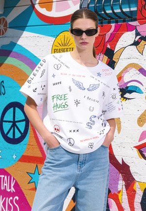 Vrouw met zwarte zonnebril en een wit t-shirt met kleurrijke handgeschreven tekst en tekeningen, staand tegen een levendige muurschildering.