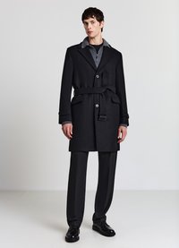 Cappotto di lana nero con colletto grigio, vita cintata, doppia fila di bottoni e due tasche frontali. Indossato sopra un dolcevita grigio strutturato e pantaloni scuri.