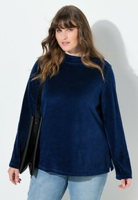 Top de terciopelo azul marino con mangas largas y aberturas laterales. Textura suave, ajuste relajado, cuello alto. Combinado con jeans azul claro y un bolso negro.