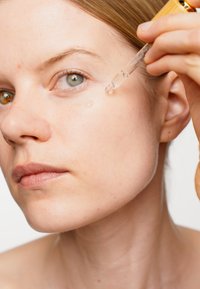 Przezroczysty serum z pipetą nałożonym w okolicy oczu. Zbliżenie na teksturę skóry, subtelny połysk i neutralne tło. Modelka o naturalnej cerze.