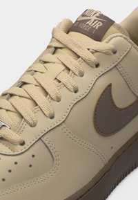 Baskets Nike Air Force 1 beige avec lacets assortis, logo swoosh marron et étiquette marron sur la languette, vue en gros plan.