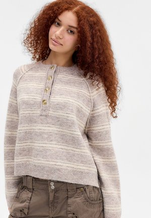 Ung kvinde med krøllet rødt hår iført en beige stribet cardigan med knapper og olivengrønne cargobukser mod en ensfarvet baggrund.