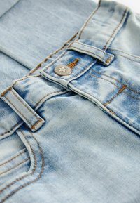 Tissu en denim bleu clair avec des coutures de poche visibles, bouton en métal avec détail en étoile et surface texturée. Présente une taille classique.