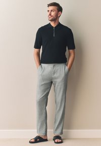 Černé polo s krátkým rukávem a zipem u krku, texturovaná látka; světle šedé kalhoty na míru; černé nazouvací sandály. Minimalistický design, volný střih.