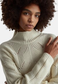 Sweter w kolorze kremowym, z dzianiny, z wysokim, prążkowanym kołnierzem. Posiada teksturowane wzory oraz otwarte detale ściegów. Dopasowane mankiety z prążkowaniem.