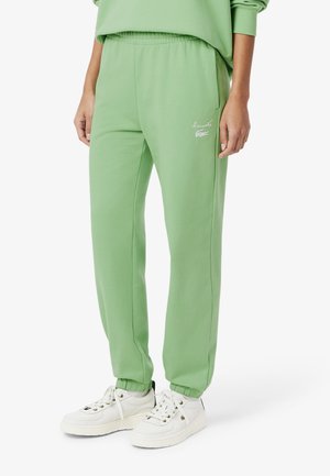 Pantaloni della tuta verdi in morbido tessuto con vita elastica, tasche laterali e un piccolo logo ricamato, abbinati a scarpe da ginnastica bianche.