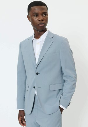 Mann in hellblauem Anzug und weißem Hemd, steht mit einer Hand in der Tasche und schaut seitlich vor einem schlichten Hintergrund.