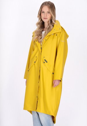 Kobieta o długich blond włosach nosi długi żółty płaszcz przeciwdeszczowy z kapturem, zamkiem błyskawicznym i zatrzaskami, stojąc na tle jednolitego białego tła.