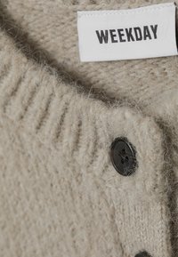 Bežani vune pleteni odjevni predmet s crnim dugmadi i bijelom etiketom ušivenom unutra koja prikazuje naziv brenda "WEEKDAY."