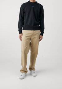 Mann in einem schwarzen Langarmpullover mit kleinem Logo, beigen geraden Hosen und weißen Sneakers, der vor einem schlichten hellen Hintergrund steht.