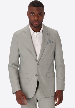Uomo che indossa un abito grigio chiaro con camicia bianca e fazzoletto da taschino a fantasia, in piedi davanti a uno sfondo bianco uniforme.