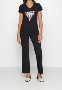 Camiseta negra de cuello en V con un diseño gráfico geométrico en color rosa, combinada con pantalones negros y zapatos de tacón cuadrado en color crema, exhibida en un modelo.