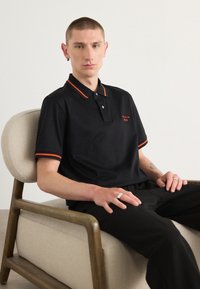 Polo noir avec des accents orange au col et aux manches. Le tissu semble lisse. Porté avec un pantalon sombre sur une chaise.