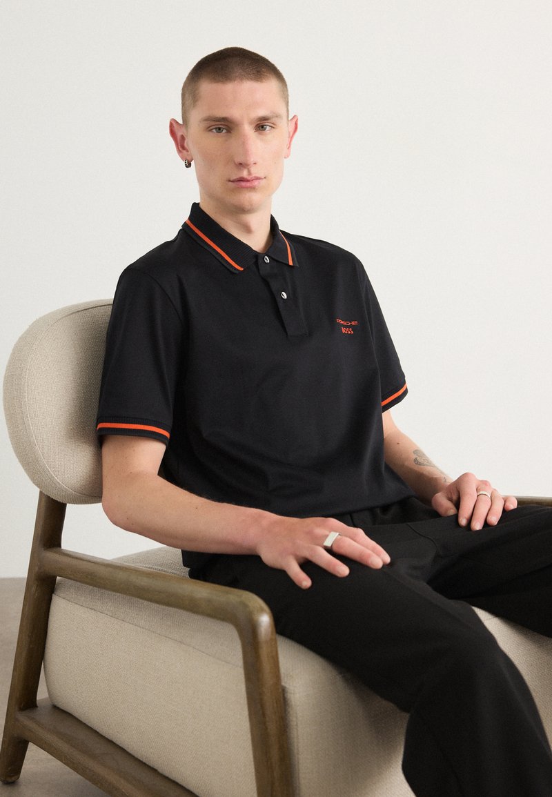 Polo noir avec des accents orange au col et aux manches. Le tissu semble lisse. Porté avec un pantalon sombre sur une chaise.
