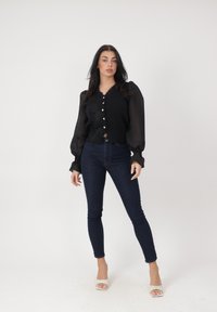 Mujer de pie que lleva una blusa negra transparente de manga larga, jeans ajustados oscuros y tacones blancos abiertos, sobre un fondo blanco liso.