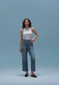 Høyt livlysende jeans med rett ben, parret med en hvit ermeløs topp. Modellen har på seg svarte loafers. En enkel blå bakgrunn.
