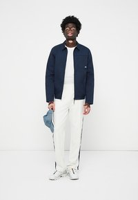 Chaqueta azul marino sobre una camisa blanca, combinada con pantalones blancos con rayas laterales y zapatillas blancas. Sosteniendo una gorra de color azul claro.