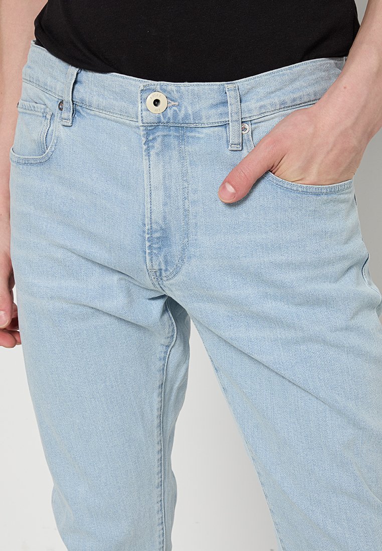 Hellblaue Jeans aus Denim an einer Person mit der rechten Hand in der Tasche und einem schwarzen Oberteil, das hineingesteckt ist, mit sichtbarem vorderem Knopf und Gürtelschlaufen.